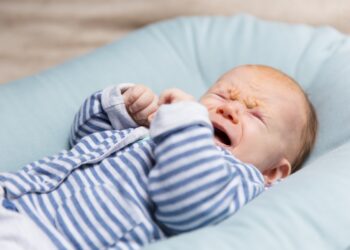 Crying red haired baby in blue and grey clothes, lying on soft pad. Portrait of cute little child in home interior. Discomfort concept
