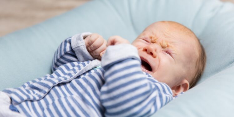 Crying red haired baby in blue and grey clothes, lying on soft pad. Portrait of cute little child in home interior. Discomfort concept