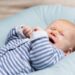 Crying red haired baby in blue and grey clothes, lying on soft pad. Portrait of cute little child in home interior. Discomfort concept