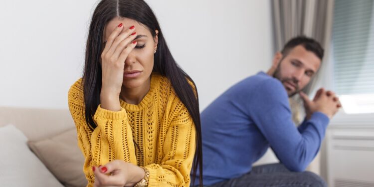 Sad pensive young girl thinking of relationships problems sitting on sofa with offended boyfriend, conflicts in marriage