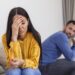 Sad pensive young girl thinking of relationships problems sitting on sofa with offended boyfriend, conflicts in marriage