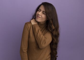young beautiful girl with closed eyes wearing brown turtleneck sweater grabbed aching shoulder isolated on purple background