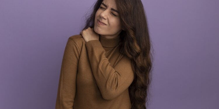 young beautiful girl with closed eyes wearing brown turtleneck sweater grabbed aching shoulder isolated on purple background