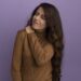 young beautiful girl with closed eyes wearing brown turtleneck sweater grabbed aching shoulder isolated on purple background