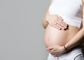 Young pregnant model in tank top touching her belly, gray background, studio, copy space, close-up