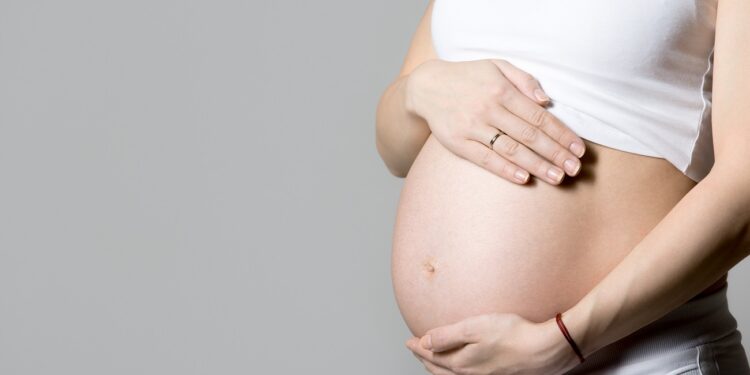 Young pregnant model in tank top touching her belly, gray background, studio, copy space, close-up