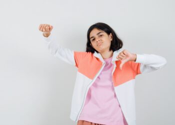 Teen girl in jacket,pink shirt showing thumb down,her fist and looking displeased , front view.