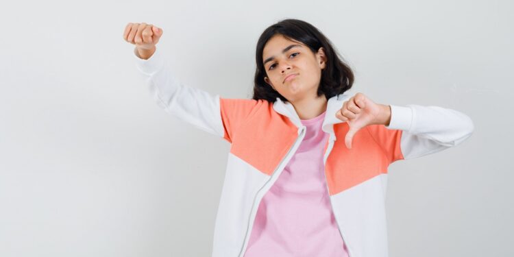 Teen girl in jacket,pink shirt showing thumb down,her fist and looking displeased , front view.