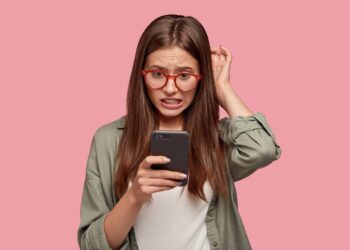 Nervous puzzled woman scratches head in bewilderment, reads bad news at internet, uses modern cell phone, wears fashionable clothes, worries aboutt something, stands in studio against pink wall