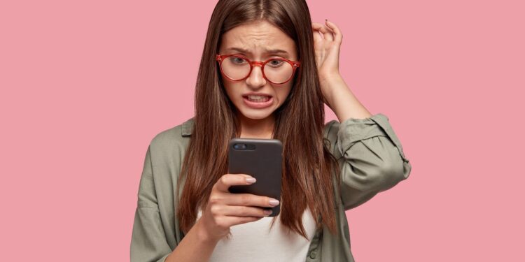 Nervous puzzled woman scratches head in bewilderment, reads bad news at internet, uses modern cell phone, wears fashionable clothes, worries aboutt something, stands in studio against pink wall