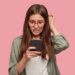 Nervous puzzled woman scratches head in bewilderment, reads bad news at internet, uses modern cell phone, wears fashionable clothes, worries aboutt something, stands in studio against pink wall