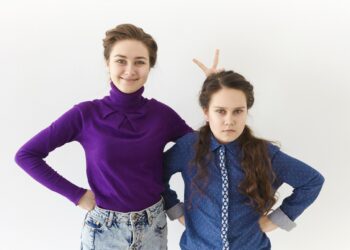 studio shot of two brunette sisters posing against white wall background: elderly girl smiling broadly making gesture, holding two fingers behind her angry sister's head for horns or rabbit ears