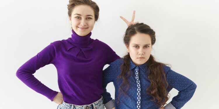 studio shot of two brunette sisters posing against white wall background: elderly girl smiling broadly making gesture, holding two fingers behind her angry sister's head for horns or rabbit ears