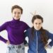 studio shot of two brunette sisters posing against white wall background: elderly girl smiling broadly making gesture, holding two fingers behind her angry sister's head for horns or rabbit ears