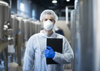 technologist with protective mask and hairnet standing at factory production line.