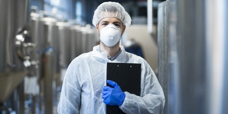 technologist with protective mask and hairnet standing at factory production line.