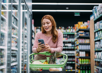 cheerful woman choosing products and using phone during grocery shopping in supermarket.