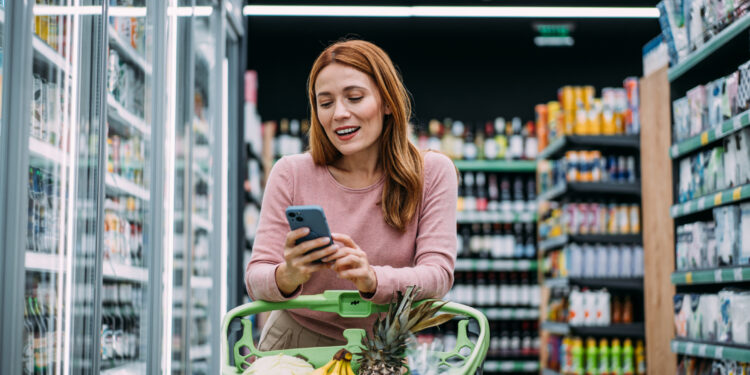 cheerful woman choosing products and using phone during grocery shopping in supermarket.