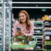 cheerful woman choosing products and using phone during grocery shopping in supermarket.