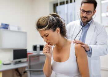 young female patient in the clinic suffered from pneumonia, she is coughing the doctor listens to the wheezing in the lungs with a stethoscope.