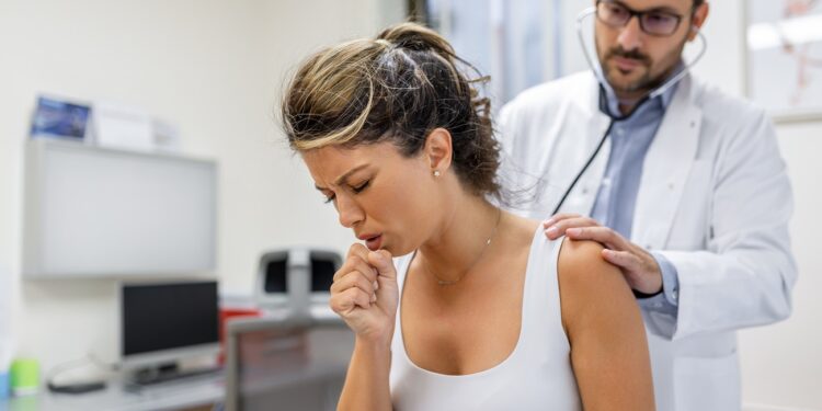 young female patient in the clinic suffered from pneumonia, she is coughing the doctor listens to the wheezing in the lungs with a stethoscope.