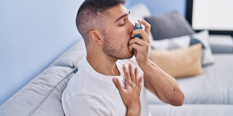 young hispanic man using inhaler sitting on sofa at home