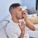 young hispanic man using inhaler sitting on sofa at home