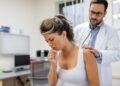 young female patient in the clinic suffered from pneumonia, she is coughing the doctor listens to the wheezing in the lungs with a stethoscope.