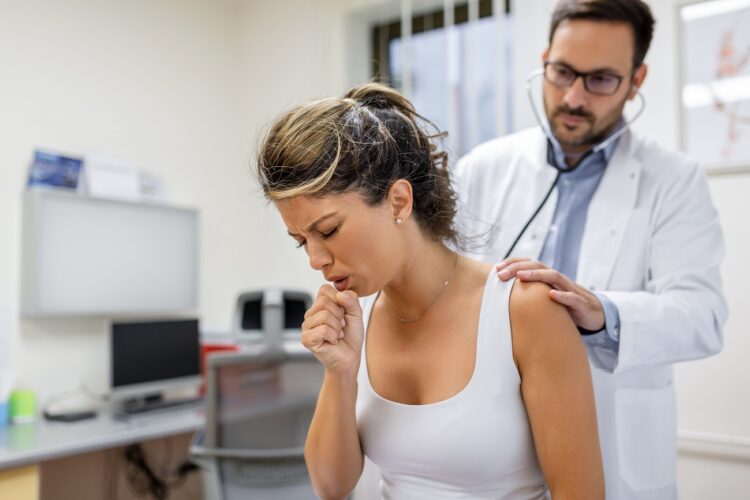 young female patient in the clinic suffered from pneumonia, she is coughing the doctor listens to the wheezing in the lungs with a stethoscope.